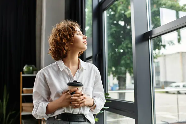 Curly-haired businesswoman stands by a large window, sipping coffee and contemplating her work. — Stock Photo