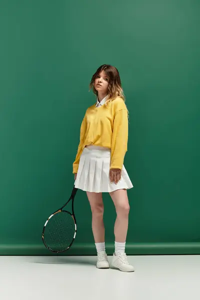 Sporty woman in a yellow sweater and white skirt holds a racket against a green backdrop. — Stock Photo