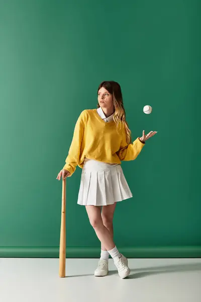 A young woman in a yellow sweater and white skirt playfully balances a baseball with a bat. — Stock Photo