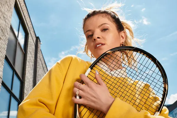 Radiant young woman in a yellow sweater poses confidently with a tennis racket, basking in sunshine. — Stock Photo