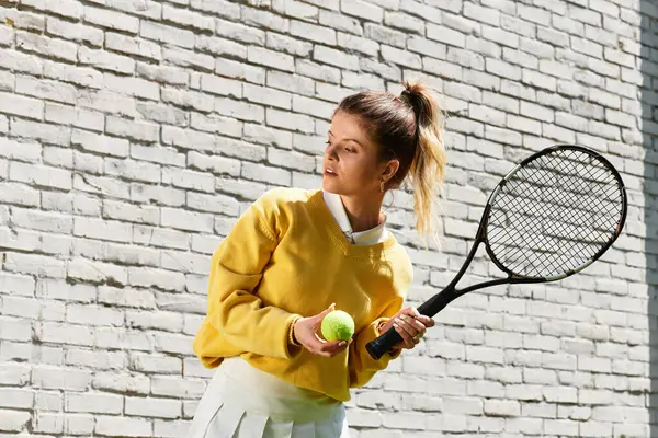 A young woman poses by a light brick wall, ready to play tennis in the summer sun. — Stock Photo
