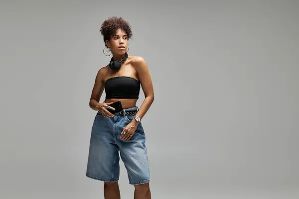 Стильна жінка з кучерявим волоссям впевнено стоїть на тлі сірого фону, тримаючи смартфон. — стокове фото