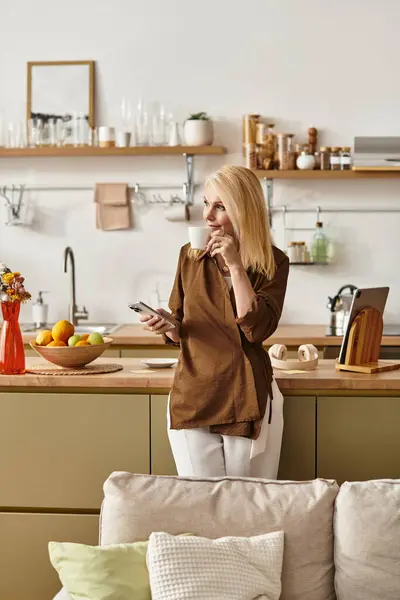 Старшая женщина с изящными чертами лица стоит задумчиво на своей стильной кухне, держа в руках телефон. — стоковое фото