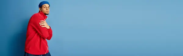 Ein junger schwarzer Mann steht selbstbewusst in roter Jacke und blauer Mütze, zeigt Stil, Banner — Stockfoto