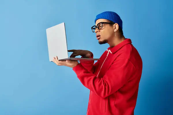 Un hombre negro joven de moda en una chaqueta roja y gorro azul utiliza su computadora portátil. - foto de stock