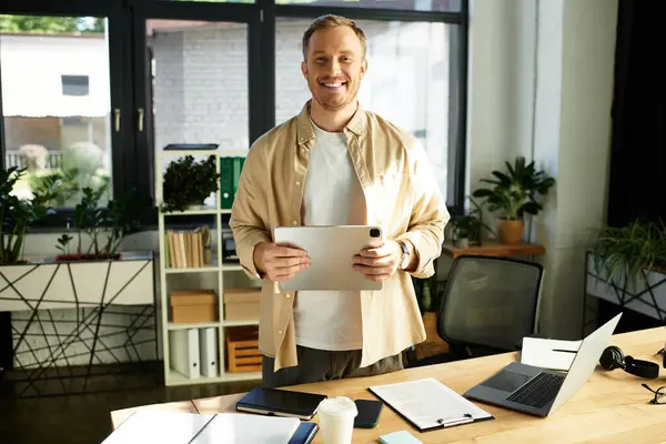 Bello giovane professionista si erge con fiducia in un ufficio luminoso mentre tiene in mano un tablet. — Foto stock