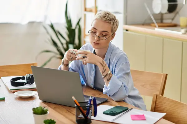 Una hermosa joven con el pelo corto se centra en su computadora portátil, sorbiendo té y trabajando en casa. - foto de stock