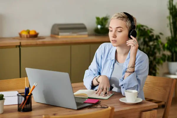 Beautiful young woman with short hair sits at a wooden table, focusing on her laptop and notepad. — Stock Photo