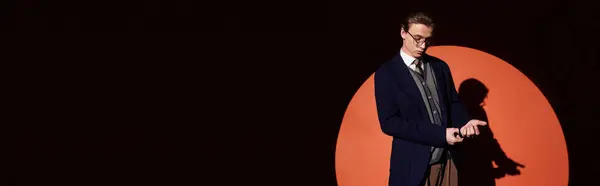 Junger gutaussehender Mann in stylischer Kleidung untersucht seine Armbanduhr mit einem dynamischen orangefarbenen Kreis dahinter. — Stockfoto