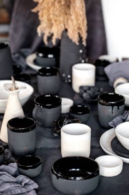 Boş el yapımı seramik sofra takımı, siyah ve beyaz kaba kaseler, kupalar, sürahi ve keten masa örtüsü üzerine vazo. Kuru kamış çiçekleri. Gün ışığı yemek odası.