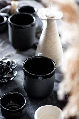 Boş el yapımı seramik sofra takımı, siyah ve beyaz kaba kaseler, kupalar, sürahi ve keten masa örtüsü üzerine vazo. Kuru kamış çiçekleri. Gün ışığı yemek odası.