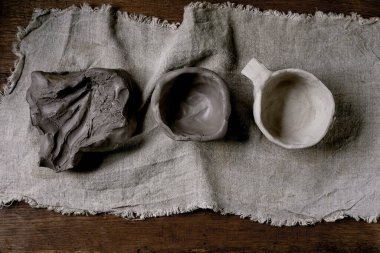 Bir parça çiğ kil, işlenmemiş çamurdan yapılmış bir bardak, seramik bardak. Seramik atölyeler ve el modelliği konsepti. Tahta masa, kaba tekstil. Üst görünüm