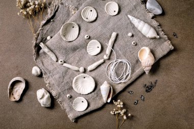 Seramik atölye fikri: kil veya porselenden yapılmış el yapımı boncuklar. Düz beyaz porselen boncuklar pürüzlü kumaş üzerinde. Küçük beyaz çiçekler, mücevher bulguları, deniz kabukları. Koyu arkaplan, üst görünüm.