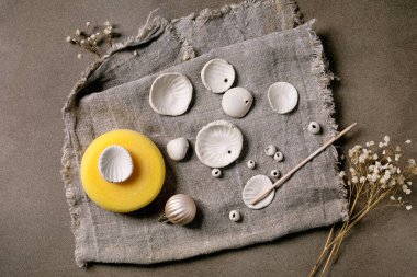 Ceramic workshop idea: handcrafted beads from clay or porcelain. Flat beads of raw white porcelain, tools on rough fabric. Small white flowers and jewelry findings. Dark background, top view. clipart