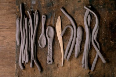 Çocuklar ve yetişkinler için çömlek atölyesi hazırlığı: bobin tekniği, el yapımı kaşıklar, koyu ahşap masadaki çömlek aletleri gibi ham kil halatları veya sosisler yığını. Üst görünüm