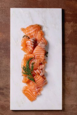 İnce dilimlenmiş somon filetosu beyaz mermer bir tahtanın üzerinde taze dereotuyla servis edilir, güzel bir yemek. Kahverengi arka plan, üst manzara. Yılın Renkleri 2025.