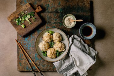 Üzerinde susam tohumu ve yeşil soğan olan gri tabakta ev yapımı Asya hamur köftesi. Soya sosu, yemek çubukları, kırsal ahşap tahta üzerinde gri peçete, bej desenli arka plan. Üst görünüm