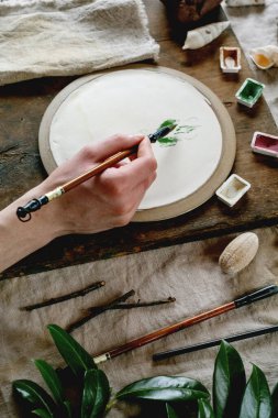 Workspace in ceramic studio, paints and brushes for underglaze painting. Young man's hands painting white plate. Branches, natural decor, linen tablecloth, daylight. Workshop idea. clipart