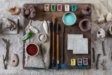 Workspace in ceramic studio, paints and brushes for underglaze painting. White tile, branches, natural decor elements, linen tablecloth. Daylight, idea for workshop. Top view clipart