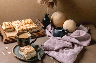 Kremalı, kremalı ve bademli ev yapımı Norveç pastası Verdens Beste Kake. Kare şeklinde kesilmiş, ahşap tepside servis ediliyor. Kahve fincanı, kaşık, süt, şişede kamış, bej masa.