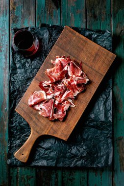 İnce dilimlenmiş İspanyol jambonu Jamon Iberico paleta bir bardak kırmızı şarapla ahşap kesme tahtasında. Eski koyu turkuaz ahşap arka plan, buruşuk siyah kağıt, üst manzara.
