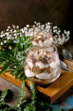 Klasik İtalyan tiramisu katmanlı tatlı, iki bardakta kakao tozu ve badem. Eski ahşap masada kaşıklarla servis edilir. Süslü beyaz bebekler nefes çiçekleri ve yeşil yapraklar.