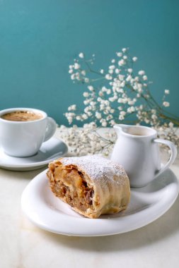 Kuru üzümlü geleneksel Avusturya elmalı turtası. Tabakta dilimlenmiş, toz şekerle toz haline getirilmiş. Beyaz mermer masa, tatlı soslu küçük sürahi, bir fincan espresso, beyaz bebek nefesli çiçekler, turkuaz duvar..