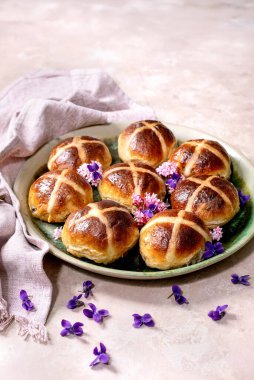 Yenilebilir çiçeklerle süslenmiş seramik tabakta geleneksel sıcak haç çörekleri. Paskalya pastası, keten peçete ve menekşe taç yapraklarıyla hafif desenli arka planda düzenlenmiştir..