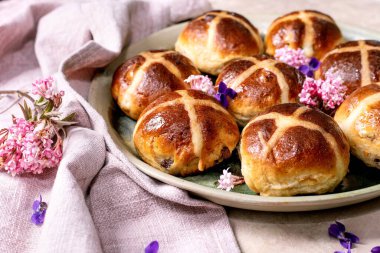 Yenilebilir çiçeklerle süslenmiş seramik tabakta geleneksel sıcak haç çörekleri. Paskalya pastası, keten peçete ve menekşe taç yapraklarıyla hafif desenli arka planda düzenlenmiştir..