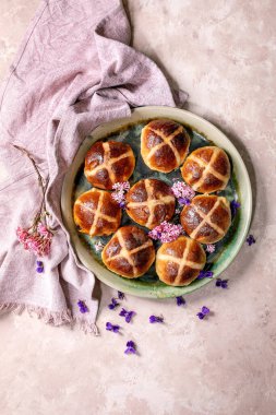 Pembe ve mor çiçeklerle süslenmiş seramik tabakta sergilenmiş sıcak haç çörekleri. Paskalya pastası, arka planda pembe keten peçeteyle. Şenlikli bahar tatili, en güzel manzarayı hazırlıyor.