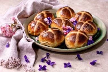 Yenilebilir çiçeklerle süslenmiş seramik tabakta geleneksel sıcak haç çörekleri. Paskalya pastası, keten peçete ve menekşe taç yapraklarıyla hafif desenli arka planda düzenlenmiştir..