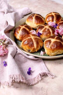 Yenilebilir çiçeklerle süslenmiş seramik tabakta geleneksel sıcak haç çörekleri. Paskalya pastası, keten peçete ve menekşe taç yapraklarıyla hafif desenli arka planda düzenlenmiştir..
