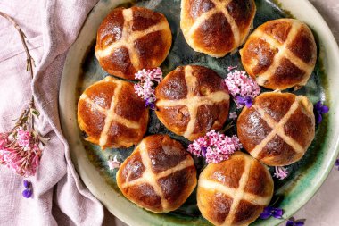 Pembe ve mor çiçeklerle süslenmiş seramik tabakta sergilenmiş sıcak haç çörekleri. Paskalya pastası, arka planda pembe keten peçeteyle. Şenlikli bahar tatili, en güzel manzarayı hazırlıyor.