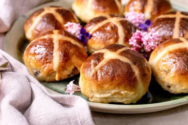 Yenilebilir çiçeklerle süslenmiş seramik tabakta geleneksel sıcak haç çörekleri. Paskalya pastası, keten peçete ve menekşe yapraklı hafif desenli arka planda düzenlenmiştir. Kapat.