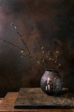 Sanatsal seramik vazoda filizlenen bahar dallarıyla minimum yaşam. Koyu kahverengi desenli arka plan. Kırsal ahşap masa. Karamsar atmosfer.