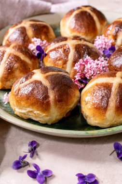 Yenilebilir çiçeklerle süslenmiş seramik tabakta geleneksel sıcak haç çörekleri. Paskalya pastası, keten peçete ve menekşe yapraklı hafif desenli arka planda düzenlenmiştir. Kapat.