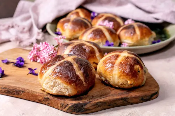 Geleneksel sıcak çörekler tahtada. Baharatlı Paskalya pastası ve bahar çiçekleriyle süslenmiş kuru meyveler. Çapraz desenli ev yapımı tatlı ekmek ruloları