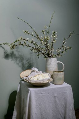 Seramik sürahide bahar çiçeği dalları, ekmek dilimli süzme peynir ve yumuşak yeşil duvara karşı beyaz masa örtüsü üzerine fincan çayı ile kırsal yaşam.