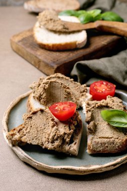 Ev yapımı ciğer ezmesi taze kiraz domatesleri ve fesleğen yapraklarıyla süslenmiş ekmek dilimlerine sürülmüş. Rustik ahşap kesme tahtası ve seramik tabak rahat aperatif sunumu oluşturur.