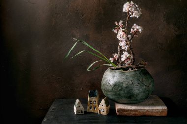 Zarif Japon tarzı hala ikebana 'da seramik tencereyle birlikte koyu kırsal arka planda kiraz dallı küçük dekoratif evler var. Taş tabanlı Wabisabi estetiği.