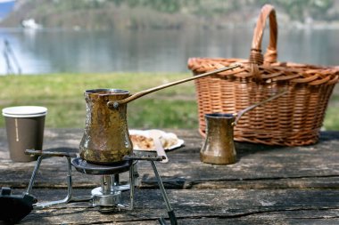 Geleneksel pirinç cezve demliği portatif soba üzerinde kahve demliyor. Hasır sepetli piknik seti ve Avusturya usulü tahta tahta masa gölü arka planlı.