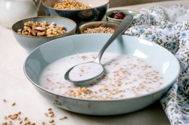 Bir kase sütlü buğday ve kaşık. Arka planda fındık taneli küçük kaseler var. Kahvaltı tezgahının yanında çiçek peçetesi. Sağlıklı sabah yemeği.