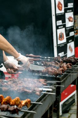 Şef eldiven giyip açık hava yiyecek pazarında et şişi pişiriyor. Arka planda menü panosu görüntüsüyle barbekü ızgarasından yükselen duman.
