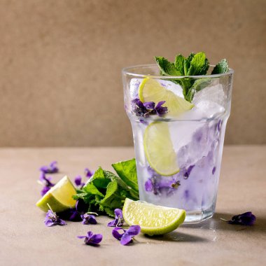 Şeffaf bir bardak çiçekli limonata kokteyli naneli, menekşe çiçekli buz ve kahverengi masada duran limon dilimleri. Soğuk yaz içeceği. Boşluğu kopyala