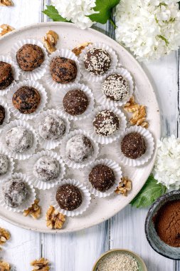 Seramik tabakta kağıt bardaklarda bulunan çeşitli ev yapımı çiğ vejetaryen topları. Farklı kaplamalar: kakao tozu şekerli susam tohumları. Ceviz ve beyaz çiçeklerle süslenmiş..