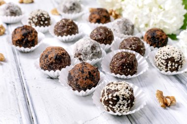 Beyaz ahşap arka planda beyaz çiçeklerle süslenmiş kağıt bardaklarda farklı malzemeler bulunan çeşitli ev yapımı çiğ vejetaryen topları.