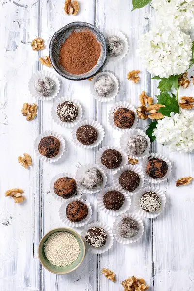 Ev yapımı çikolatalı çiğ vejetaryen toplar kağıt bardaklarda mantar kakao tozu susam tohumu ceviz ve kırsal beyaz ahşap arka planda beyaz ortanca çiçekleri. Üst manzara sağlıklı tatlı.