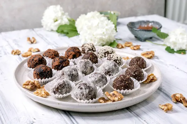 Beyaz seramik tabakta farklı malzemelerle ev yapımı çiğ vejetaryen topları. Kakao tozu ve susam tohumuyla kaplanmış. Ceviz ve beyaz çiçeklerle süslenmiş..
