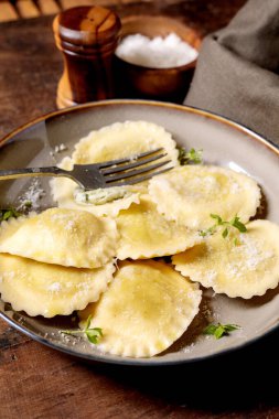 Seramik tabakta servis edilen peynirle dolu taze ev yapımı ravioli makarna otlarla süslenmiş çatal ve sade ahşap masada parmesan peyniri.