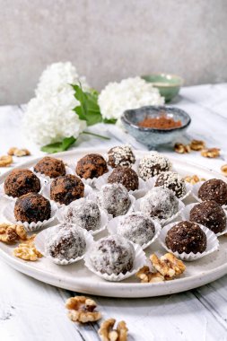 Çeşitli ev yapımı çikolatalı çiğ vejetaryen topları beyaz tabakta farklı malzemelerle. Kakao tozu cevizleri etrafa saçılmış. Beyaz çiçek arkaplanlı küçük kaseler..